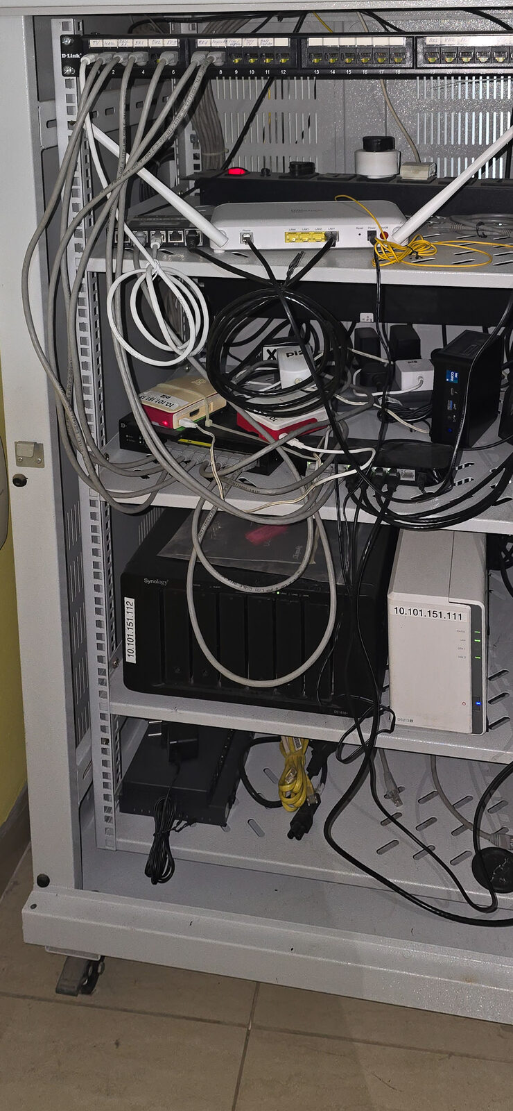 Photograph of the homelab rack: patch panel, PoE and non-PoE network switches, ISP router and APs, Raspberry Pis, an Intel NUC, and Synology NAS units, with cabling running between them.